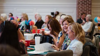 Women are seated around tables and on benches filling Fellowship Hall at St. Luke's United Methodist Church, smiling as they attend a seminar for the Women's Retreat.