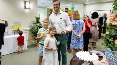 A father and his two children hold drinks and snacks as they pose for the camera in St. Luke's Gathering Room, amidst a crowd of fellow worshippers.