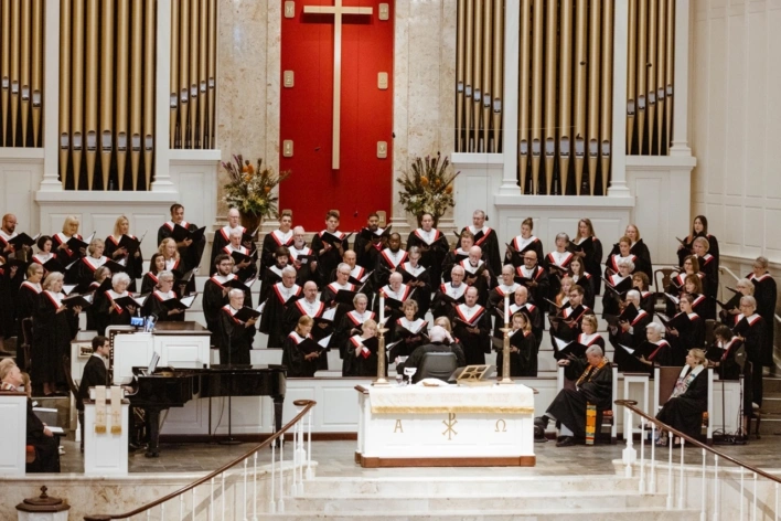 A choir of around 50 people stands under a large cross in the St. Luke's Sanctuary, directed by a man in a wheelchair.