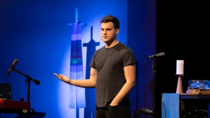 Rev. Colin Bagby stands on stage in branches' worship space, wearing a t-shirt and jeans in a more modern worship setting.